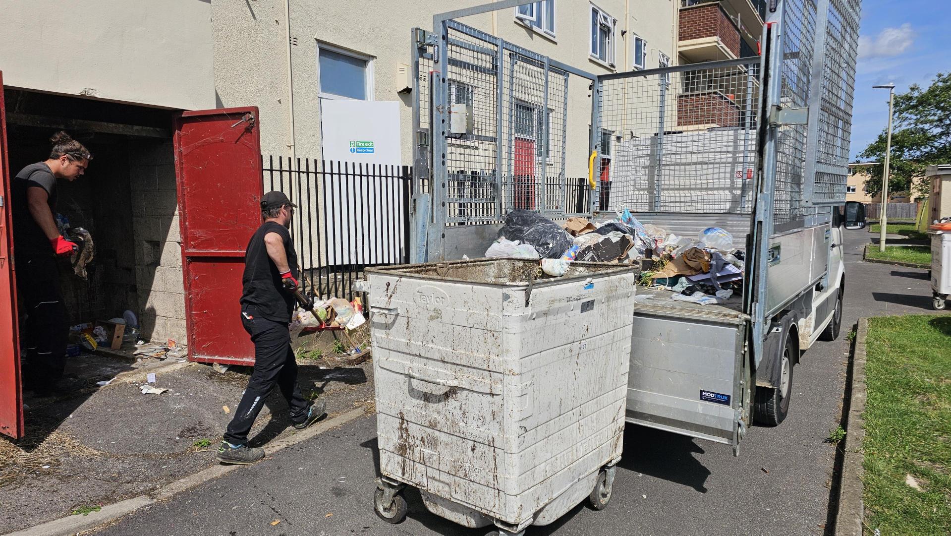 A GCH colleague shovelling rubbish on to a truck with a large wheeled bin in the foreground